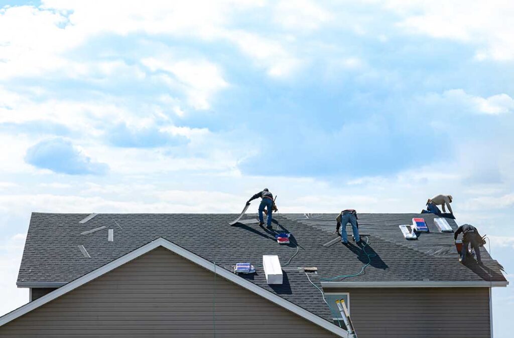 Person in a safety harness carefully walking across a dry shingle roof beside a secured ladder.