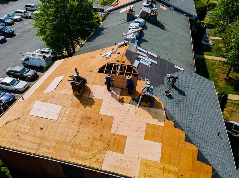 Workers remove old shingles from a house roof while protective tarps cover the landscaping below.